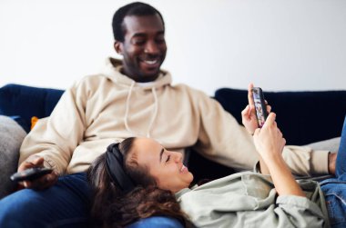 Genç, etnik kökenli bir çift evdeki kanepede televizyon izleyen bir erkekle dinlenirken, bir kadın kucağında uzanıp cep telefonundan sosyal medyayı kontrol ediyor.