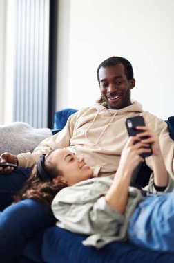 Genç, etnik kökenli bir çift evdeki kanepede televizyon izleyen bir erkekle dinlenirken, bir kadın kucağında uzanıp cep telefonundan sosyal medyayı kontrol ediyor.