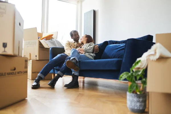 Young mixed ethnicity couple celebrating moving day in new home sitting on sofa in lounge surrounded by removal boxes together