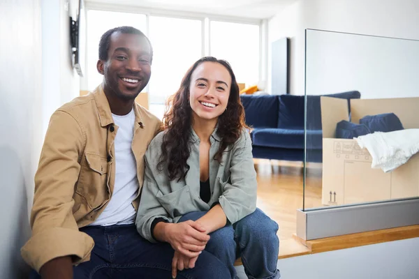 Portrait of young mixed ethnicity couple celebrating moving day in lounge of new home sitting on floor surrounded by removal boxes together