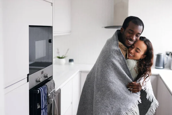 Loving young mixed ethnicity couple cuddling whilst standing in kitchen wrapped in cosy blanket together