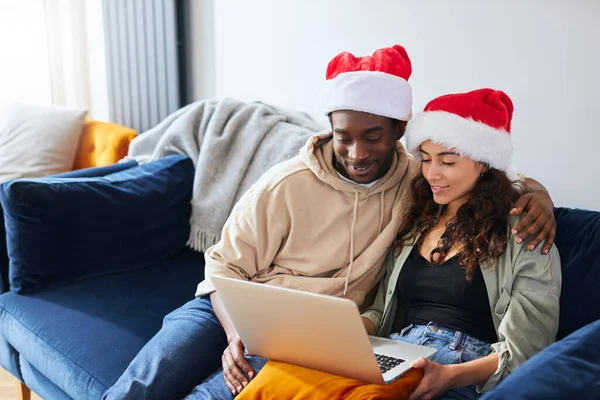 Young mixed ethnicity couple wearing santa hats sit side by side on sofa at home shopping for christmas presents online using laptop