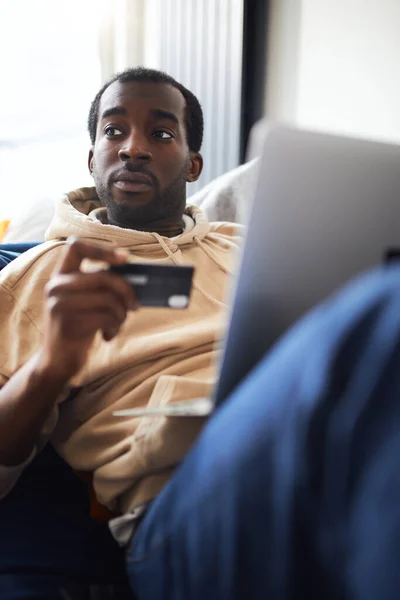 若い混合民族の男の側のビューは オンラインショップにクレジットカードを使用してノートパソコンのコンピュータと自宅でソファに横たわっていた — ストック写真