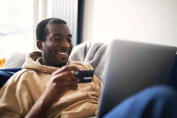 Profile view of young mixed ethnicity man lying on sofa at home with laptop computer using credit card to shop online