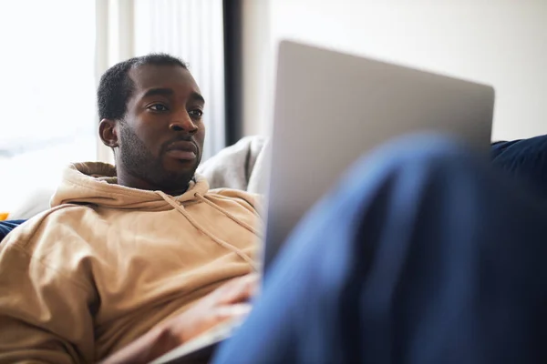 自宅やオンラインショップから動作するようにラップトップコンピュータを使用して自宅でソファの上に横たわる若い混合民族の男のプロフィールビュー — ストック写真