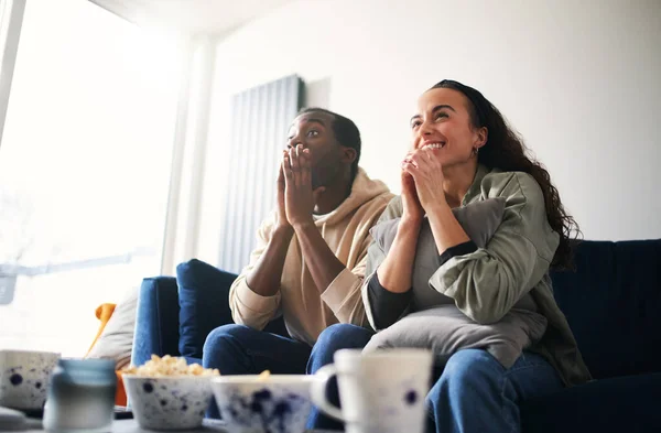 Excited and tense young mixed ethnicity couple sitting on sofa at home watching or streaming sports event on tv together