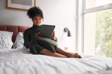 Evdeki yatakta dinlenen kadın dijital tablette TV programı okuyor ya da yayınlıyor.