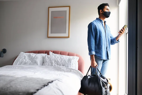 Butik otelde maskeli bir adam cep telefonu uygulamasından taksi çağırılmasını bekliyor.