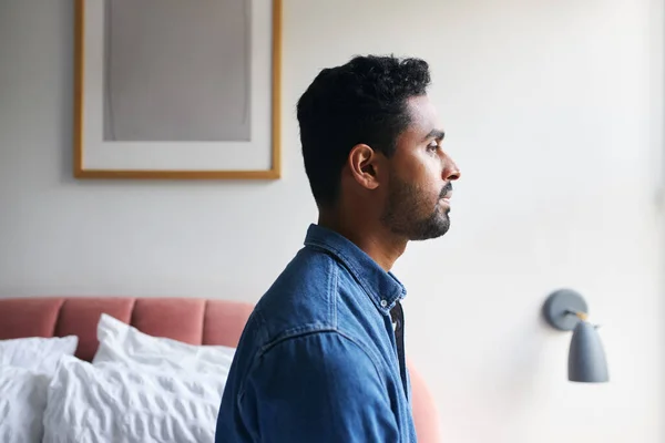 Unhappy young man with mental health issues sitting on edge of bed and staring out of window shot in profile