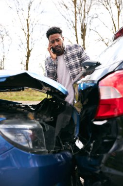 Young man standing by damaged car after traffic accident reporting incident to insurance company using mobile phone clipart