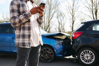 Close up of man standing by damaged car after traffic accident reporting incident to insurance company using mobile phone clipart