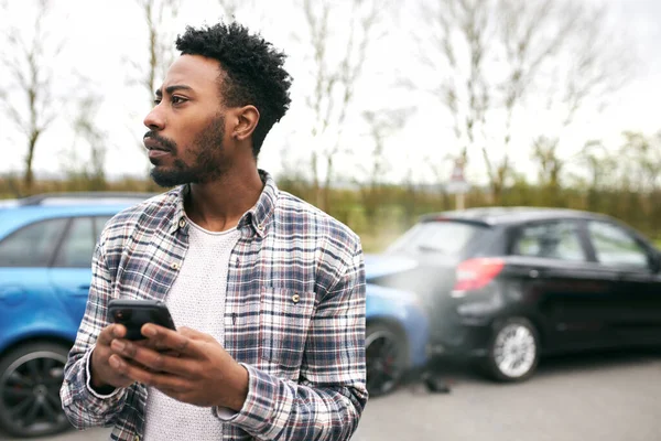 交通事故の後に破損した車で立っている若い男は 携帯電話を使用して復旧会社を呼び出します — ストック写真