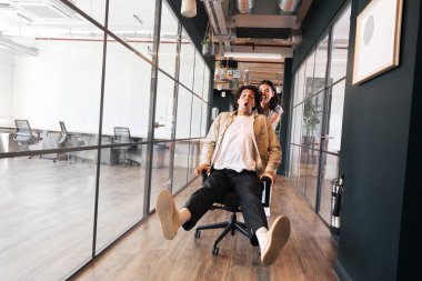 Kadın olarak eğlenen genç çift, erkeği modern açık ofis sandalyesinde yürümeye zorluyor.