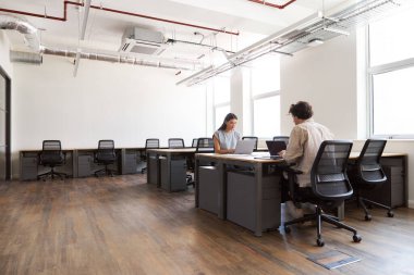 Modern açık planlama ofisindeki masalarda dizüstü bilgisayarlar üzerinde çalışan sıradan giyinmiş genç adam ve kadın.