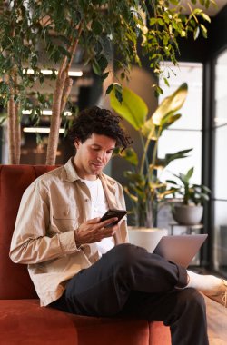 Sıradan giyimli genç adam cep telefonunu kontrol ediyor modern açık planlı ofisin oturma alanında bilgisayarla çalışıyor.