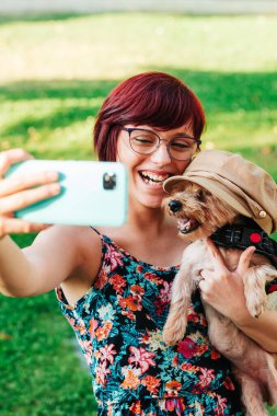 Yorkshire 'lı köpeğiyle cep telefonuyla selfie çeken mutlu ve komik genç bir kadının dikey görüntüsü..