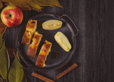 Elmalı turta demir kasede, sonbahar yaprakları solda siyah masada ve yazı için boşluk sağda, yakın çekim üst görünümde..