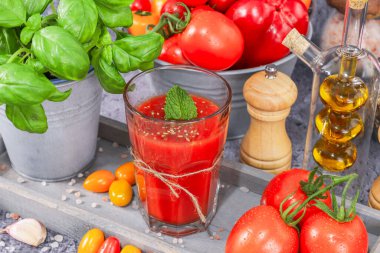 Bir bardakta domates suyu, fesleğen, domates, zeytinyağı ve baharatlar gri bir tepside taş bir arka plana çapraz yerleştirilmiş..