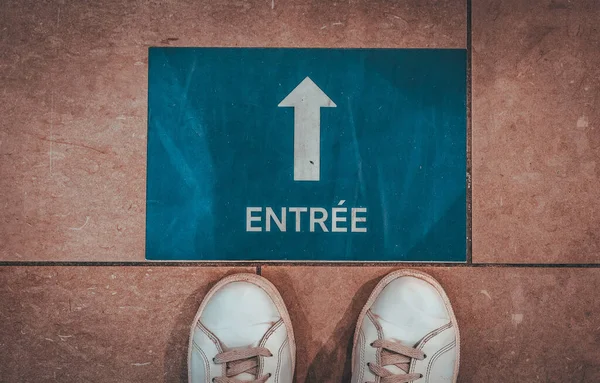 Forward arrow labeled in French: Entrance, in blue square with white sneakers on brown tiles, top view.