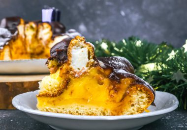 Bir dilim çikolatalı pasta, profiterollü krema, beyaz bir fincan tabağında krema, koyu bir arka planda noel dekoru, yakın plan yan görüş..