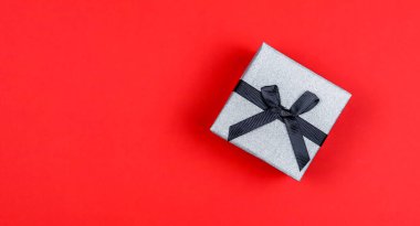 Gri bir hediye kutusu. Sağında siyah kurdele var. Kırmızı arka planda..