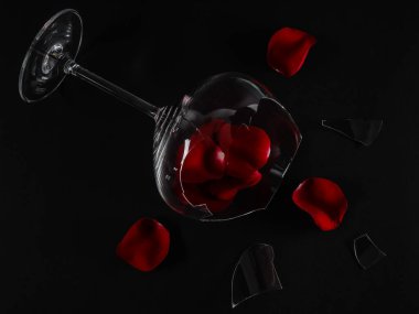 A broken glass and rose petals with shards lie diagonally on a black background, close-up top view.