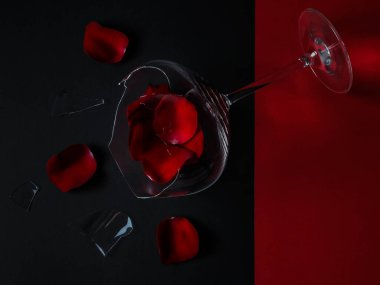Broken glass and rose petals lie diagonally against a red and black background, close-up top view.