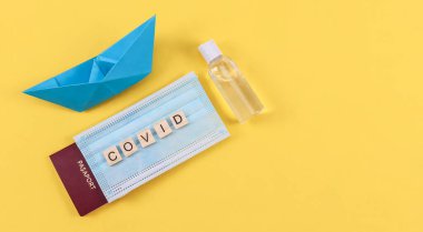 Maskeli pasaport ve solunda dezenfektan olan kağıt tekne sarı arka planda sağda yazı için boşluk, üstte görünüm, yakın çekim..