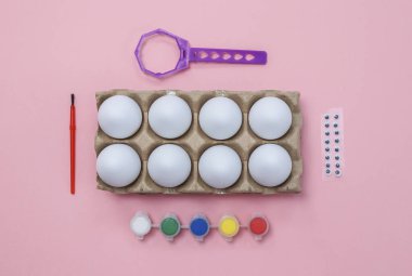 Sekiz beyaz yumurta tepsisi, boya, fırça ve göz, pembe arka planda yatar, üst taraf yakın plan.