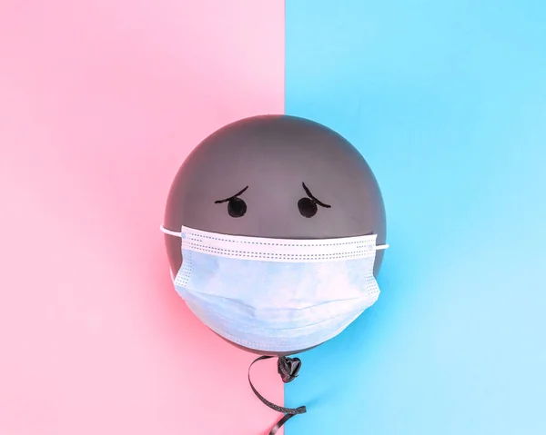 A gray balloon with a mask and a sad emotion lies in the middle on a pink-blue background, a close-up top view.