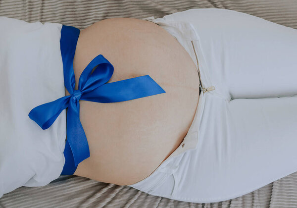 Blue ribbon tied in a bow on the belly of a pregnant woman, close-up top view.