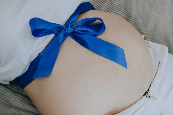 Belly of a pregnant woman tied with a blue ribbon lying on her back in bed, top view close-up.