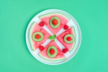 Watermelon with berries and fruits in a plate on a green background, top view close-up.