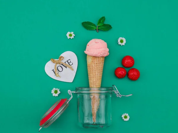 Homemade strawberry ice cream in a jar with mint, flowers and heart on a green background, top view close-up.