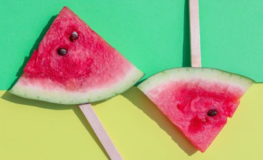 Çubuklardaki karpuz dilimleri sarı-yeşil bir zemin üzerinde uzanır..