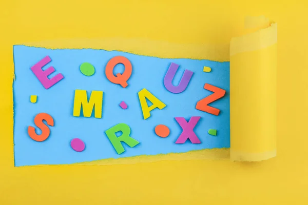 Colored letters of the alphabet on a blue background in a yellow frame of torn paper with a curl, top view close-up.