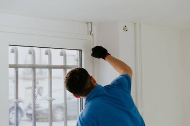 Mavi tişörtlü, koyu renk sakallı, genç bir beyaz adam pencerenin önünde dikiliyor ve pencereyi beyaz bir fırçayla boyuyor..