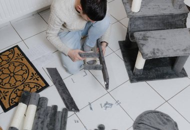 Yeni bir kedi evinin parçalarını birleştiren esmer, beyaz bir adamın portresi oturma odasında yerde otururken, üst taraf manzaralı yakın plan. Kedi mobilyası montajı ve kazıma direkleri kavramı.