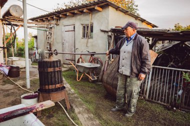 Kirli iş kıyafetleri içinde, şarap makinesinde, levyeyle üzüm suyu sıkan, kırsal bir arka bahçede sonbahar günü yan duran yaşlı beyaz bir adamın tam boy portresi. Geniş açı. Kavram: Geleneksel şarap yapımı.