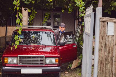 Yaşlı bir adam ve kadın, bulutlu bir sonbahar gününde üzüm hasadı taşımak için köy avlusunda park etmiş kırmızı bir arabaya binmeye hazırlanıyorlar. Yan görüntüyü yakından çek. Konsept: Hasat zamanı, geleneksel şarap yapımı, ulaşım hizmetleri.