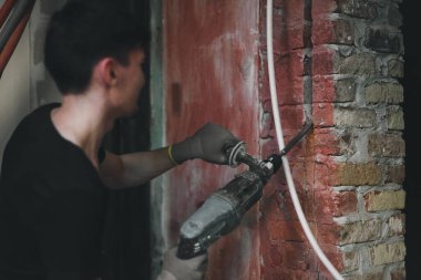 Siyah üniformalı, tanınan genç bir beyaz adam, seçici bir odak noktasıyla merdiven üzerinde dikilirken büyük bir tornavidayla tuğla duvarda bir delik açıyor. Ev yenileme konsepti, inşaat işi.
