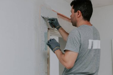 Gri üniformalı ve eldivenli genç beyaz tenli bir esmer, inşaat ızgarasını beyaz macunla kaplıyor, kapının önündeki merdiven yarı yarıya dönük, aşağıdan seçici bir odak ile yakın çekimde. Ev yenileme konsepti