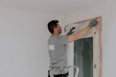 Gri tişörtlü genç beyaz bir adam merdivenin üzerinde duruyor ve kapı eşiğinin üstündeki duvara dar inşaat ızgarası yerleştiriyor. Bahar günü odak noktası seçici bir şekilde yan tarafından orta mesafeden vurulmuş. Macun ve inşaat işleri konsepti.