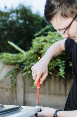 Dikey portre genç Kafkasyalı yakışıklı adam tornavidayla düzüşüyor. Yazın arka bahçede metal barakanın çatısını kuruyor. Yakın plan. Kavramsal metal dökümü montajı,