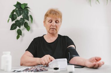 Son sınıf güzel bir kadının yatay portresi. Tansiyonunu tonometreyle ölçen gri masada ayakta oturan ve gündüz vakti odada oturan bir kadın. Alt tarafı orta ölçekli. Teknoloji ve emeklilik yaşam tarzı kullanma kavramı.