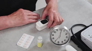 Siyah tişörtlü yaşlı bir kadın kalp atış hızı monitörü alıyor ve nabzı ölçüyor. Gündüz vakti odasındaki tahta masada oturuyor. Üst tarafı yavaş çekimle kapatıyor. Teknoloji kullanımı zamanı, emeklilik yaşam tarzı kavramı.