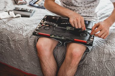 Genç, beyaz, tanınmayan bir adam laptopun devre kartındaki kabloları düzeltirken aynı zamanda odanın üst tarafındaki yatakta oturuyor. Adım adım dizüstü bilgisayar tamiri ve termal yapıştırıcı değişimi kavramı.