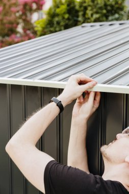 Omuz hizasında, siyah tişörtlü ve gözlüklü beyaz bir adamın dikey portresi. Yazın arka bahçede metal barakanın çatısına manuel olarak cıvatalar takıyor. Yan görüş çok yakın. Teknolojiyi kullanarak metal döküntülerini birleştir.