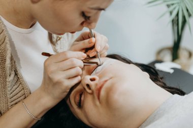 Genç ve güzel bir beyaz kadın kozmetik uzmanının portresi. Yapay kirpikler yapıştırıyor. Kadın bir müşterinin sol gözüne iki cımbız yapıştırıyor. Kirpik uzantısı