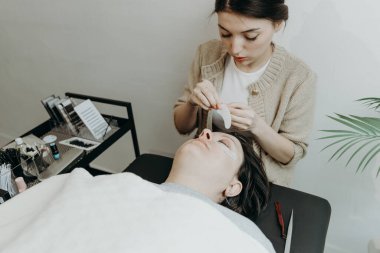 Beyaz tenli güzel bir kozmetik uzmanı kızın portresi, bir bahar günü ofiste kozmetoloji masasında uzanan bir bayanın sol gözkapağının alt göz kapağı için kağıt yapıştırmasını soyar. Kirpik.